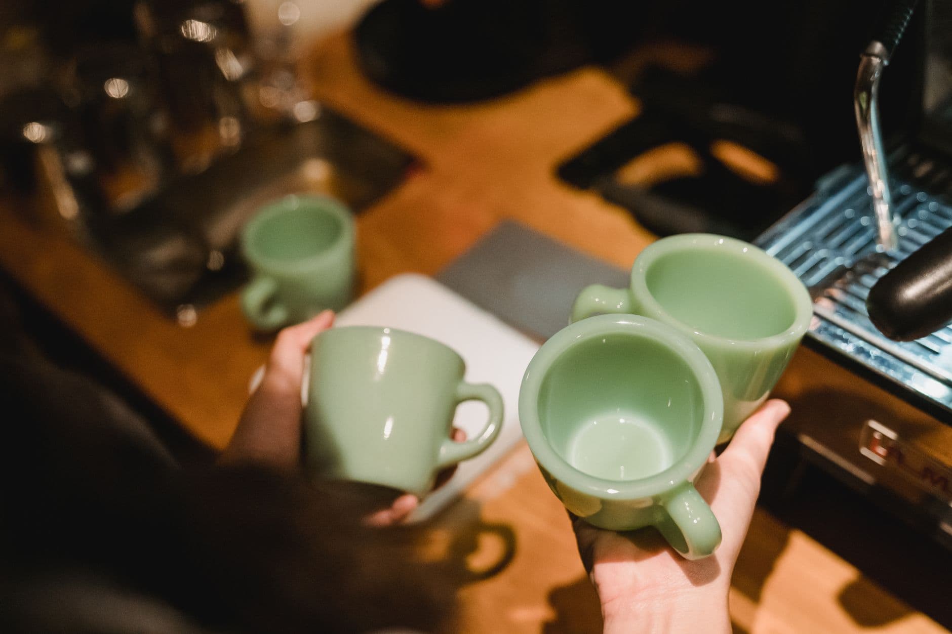 Hands holding green coffee cups near espresso machine in a café setting.