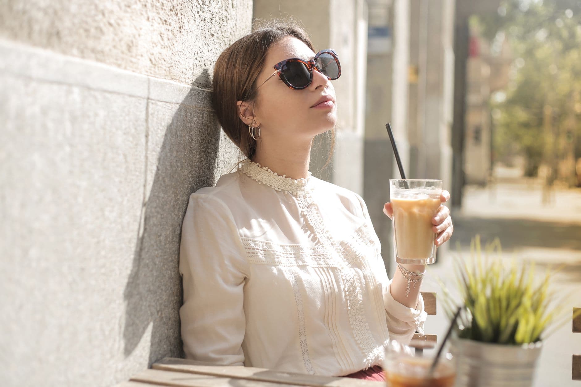 A serene woman in sunglasses enjoys a cold coffee drink in a sunny outdoor cafe setting.