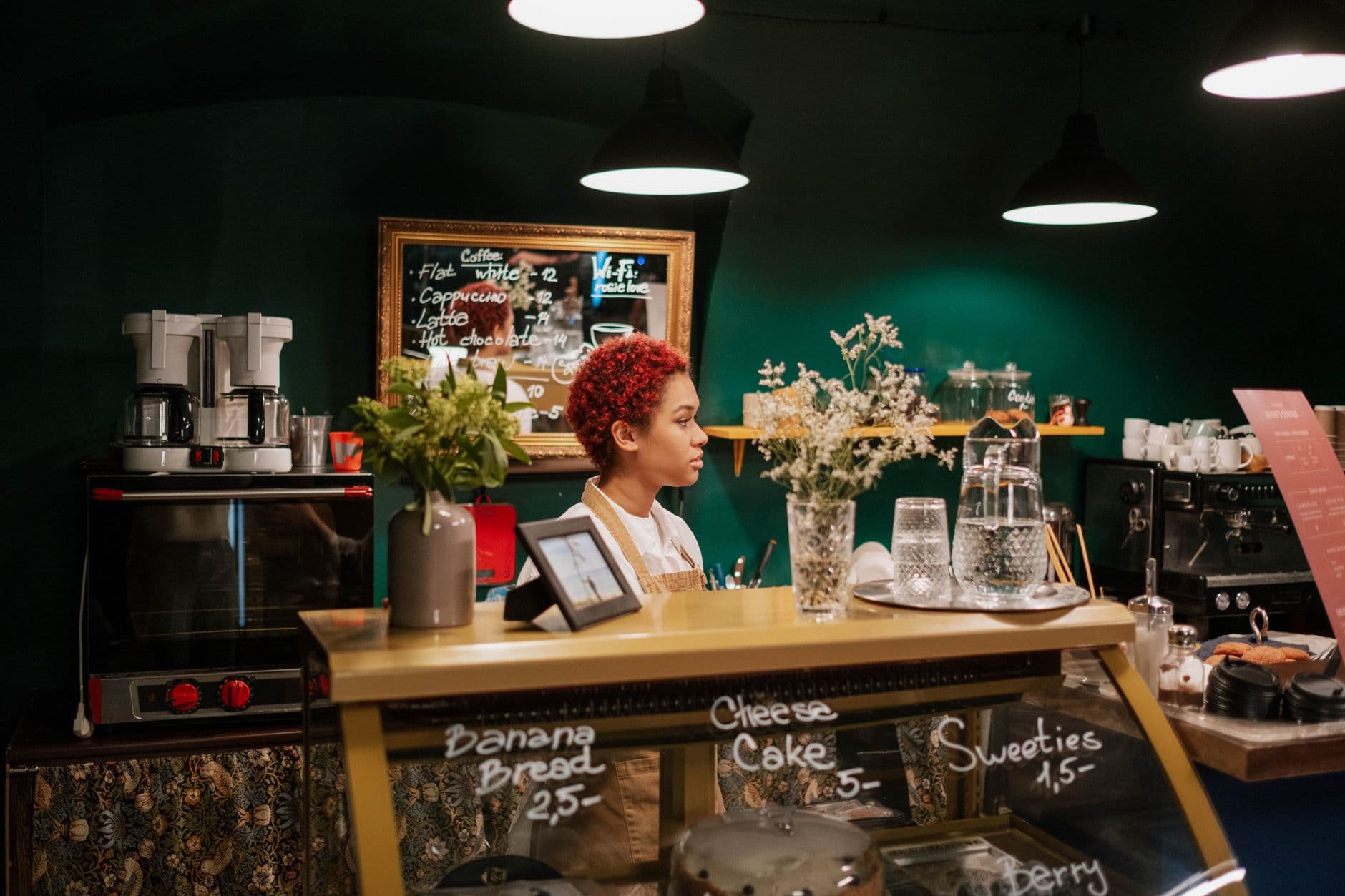 A barista stands at a café counter surrounded by pastries and coffee machines.