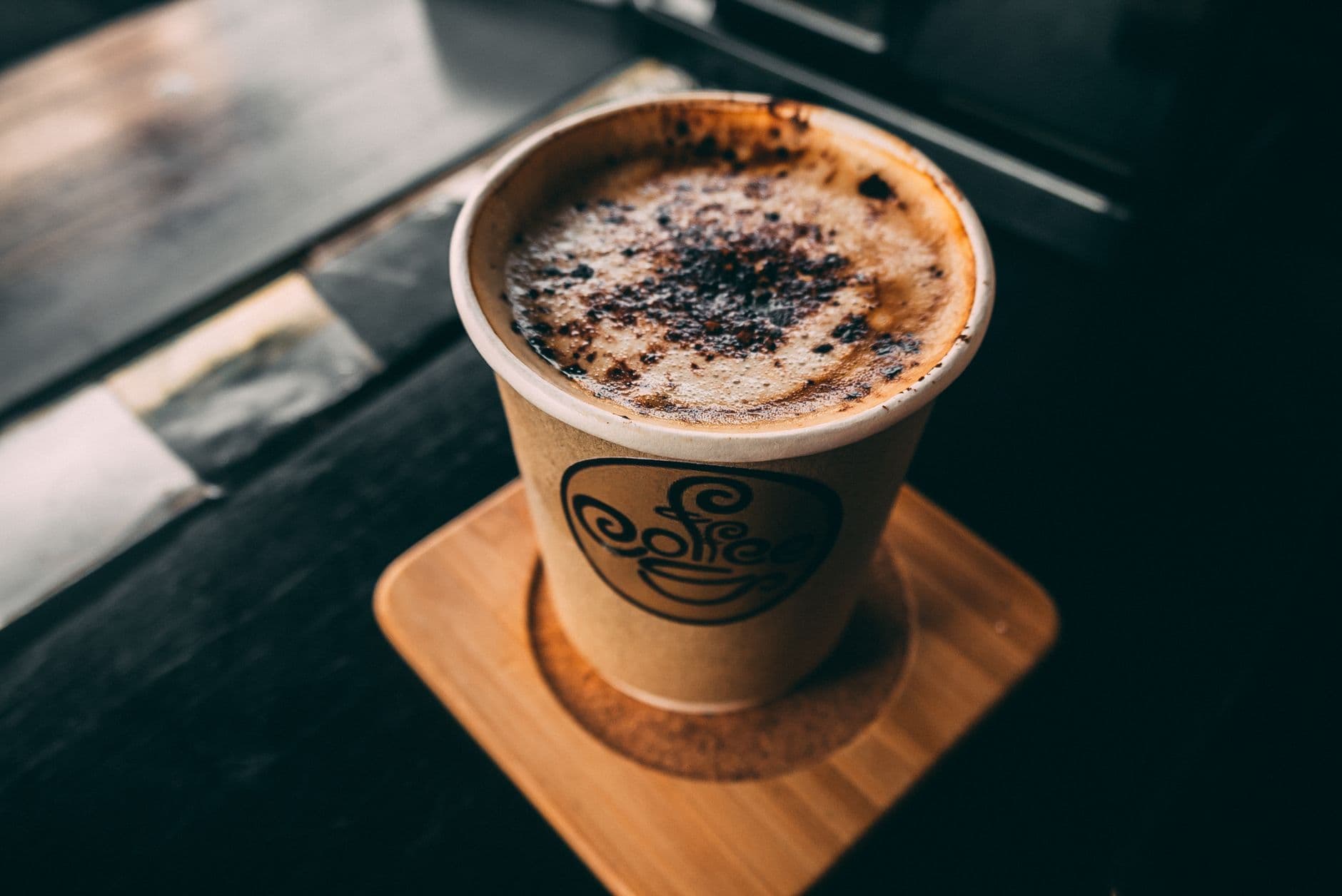 A close-up view of a cappuccino in a paper cup with foam and chocolate sprinkles on top.