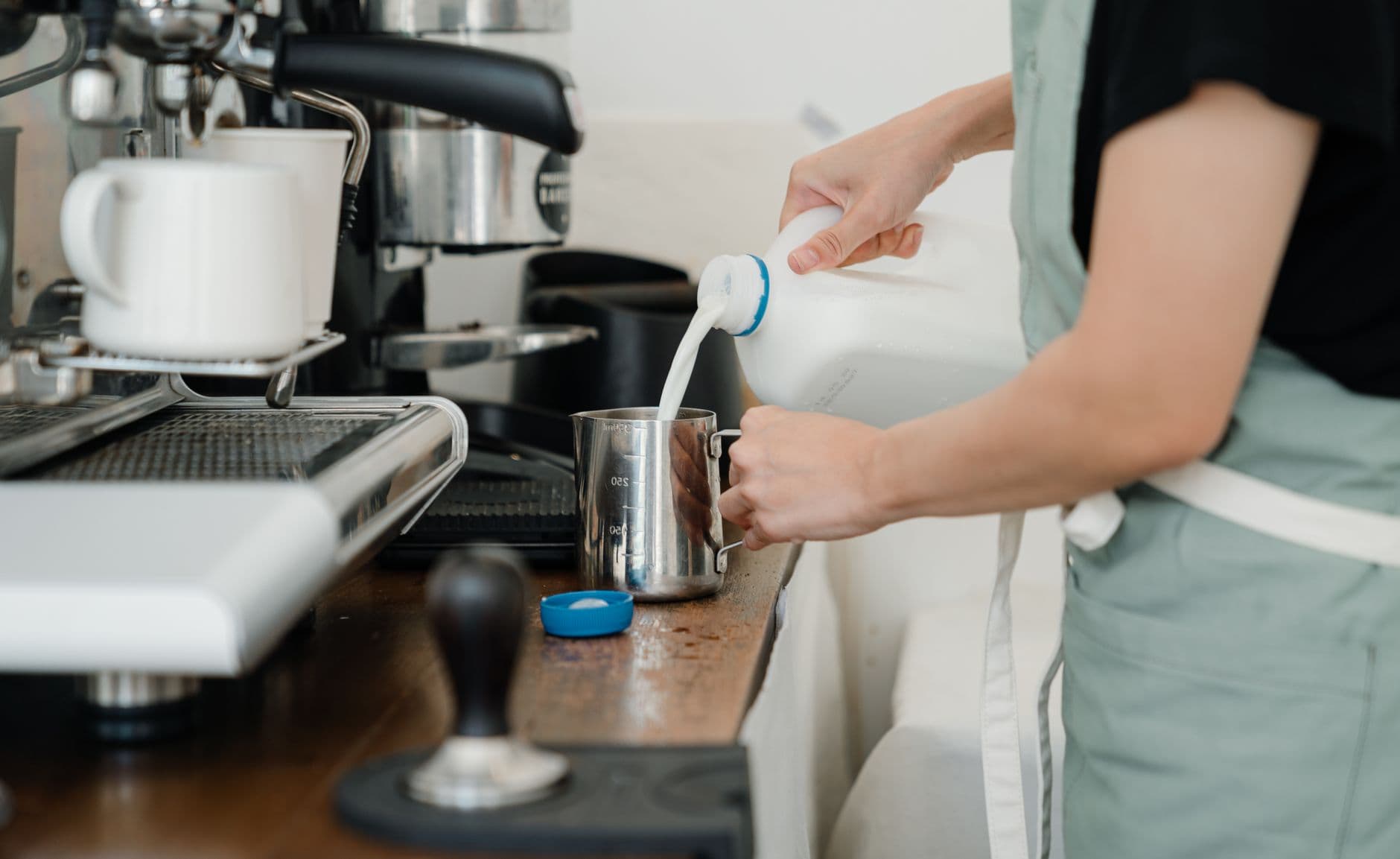Crop anonymous young female in apron pouring milk into metal pot while preparing delicious latte in modern coffee shop