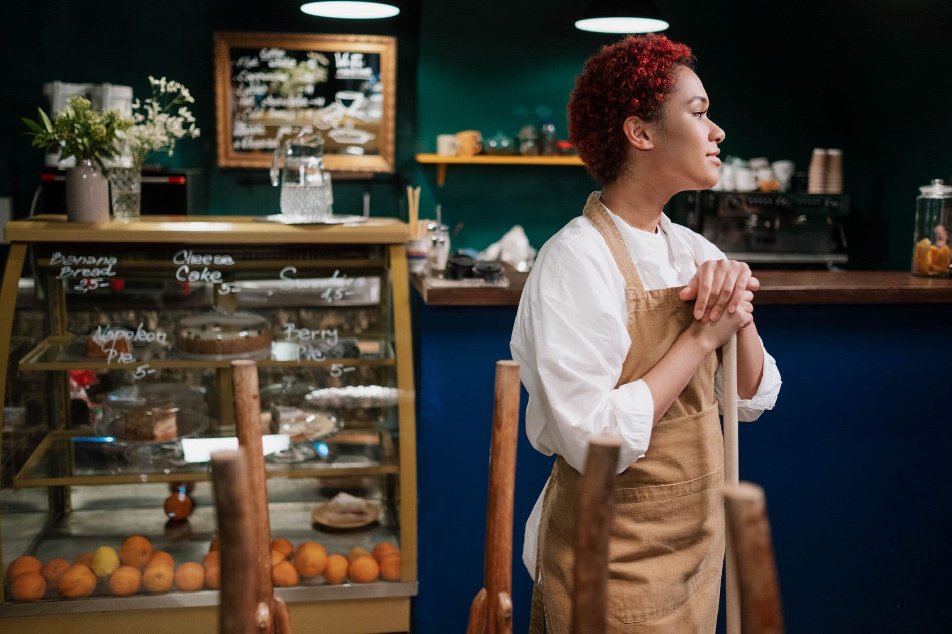 Waitress in a café wearing an apron stands thoughtfully by the counter with desserts on display.