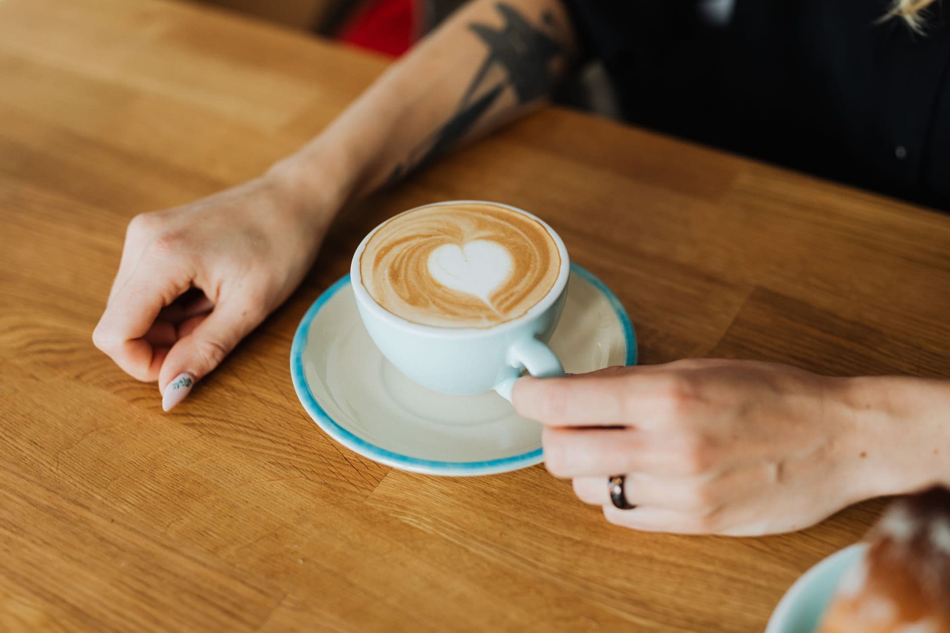 A close-up of a cappuccino with heart latte art held by tattooed hands.