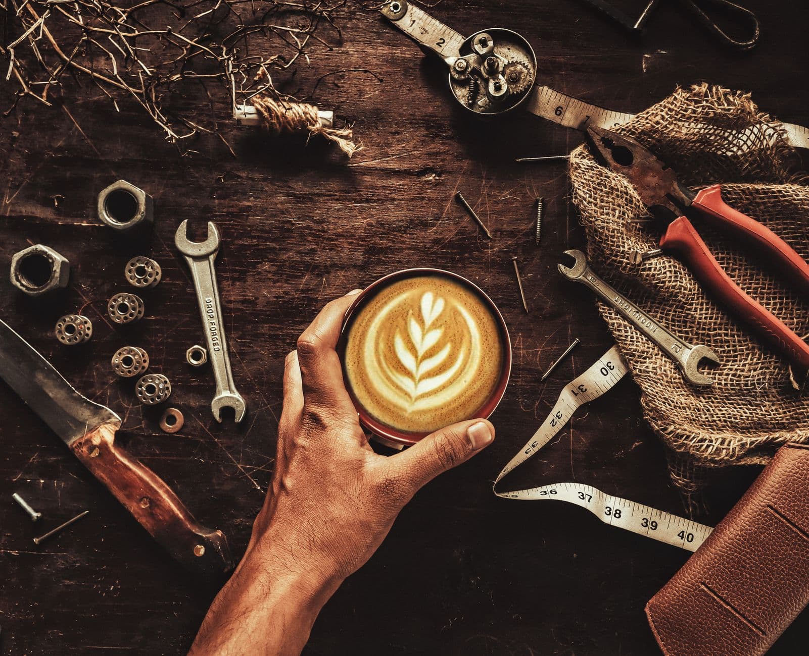 A creative flat lay of a hand holding a latte amidst various tools on a wooden surface.