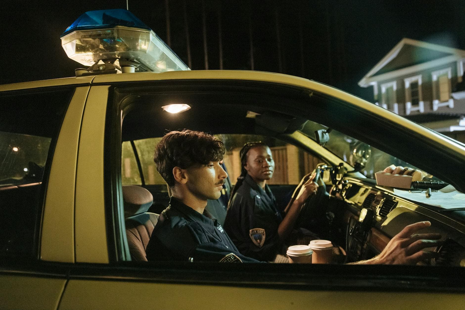 Police officers sit in a patrol car at night, holding coffee cups, ready for duty.