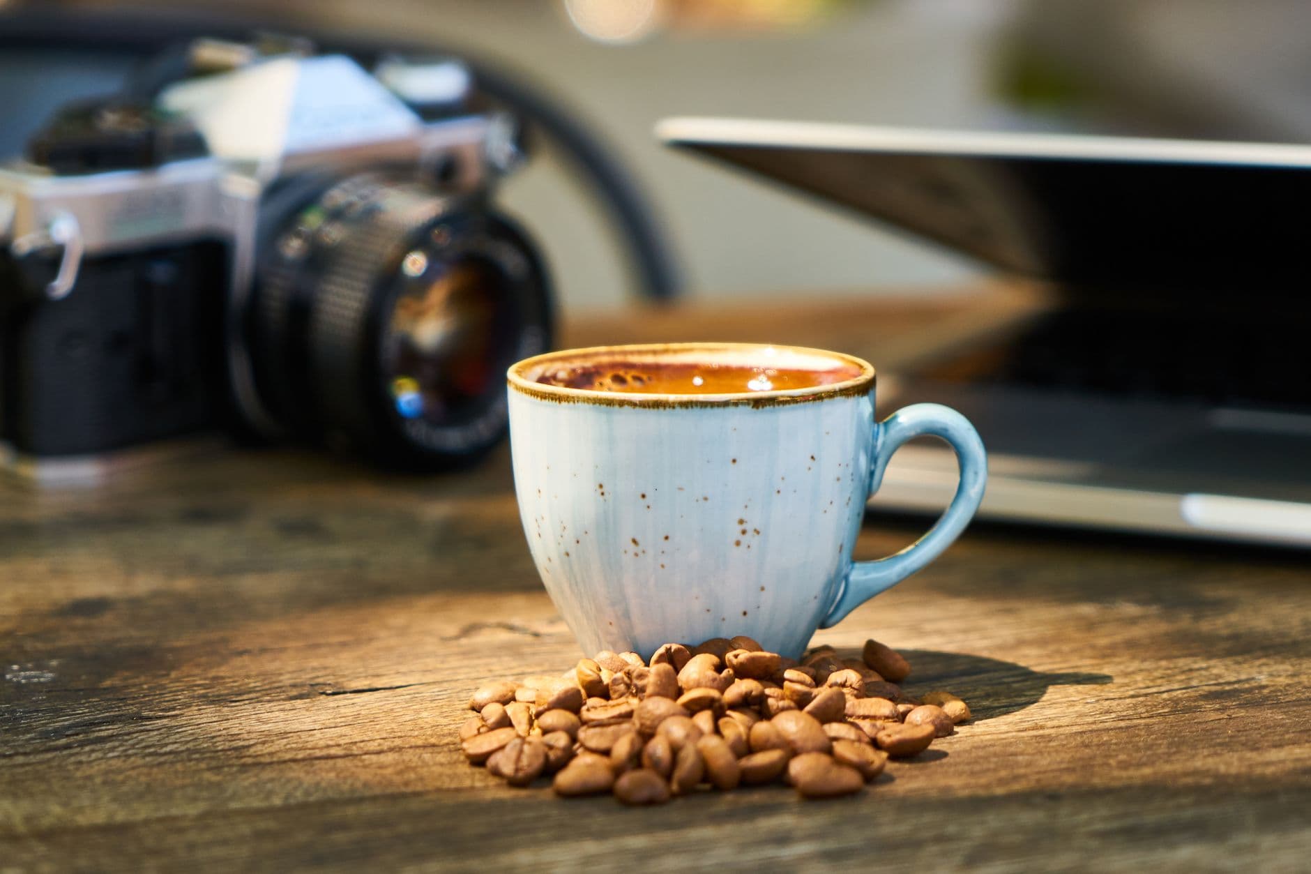 A rustic coffee cup surrounded by beans on a wooden table with a camera and laptop in the background.