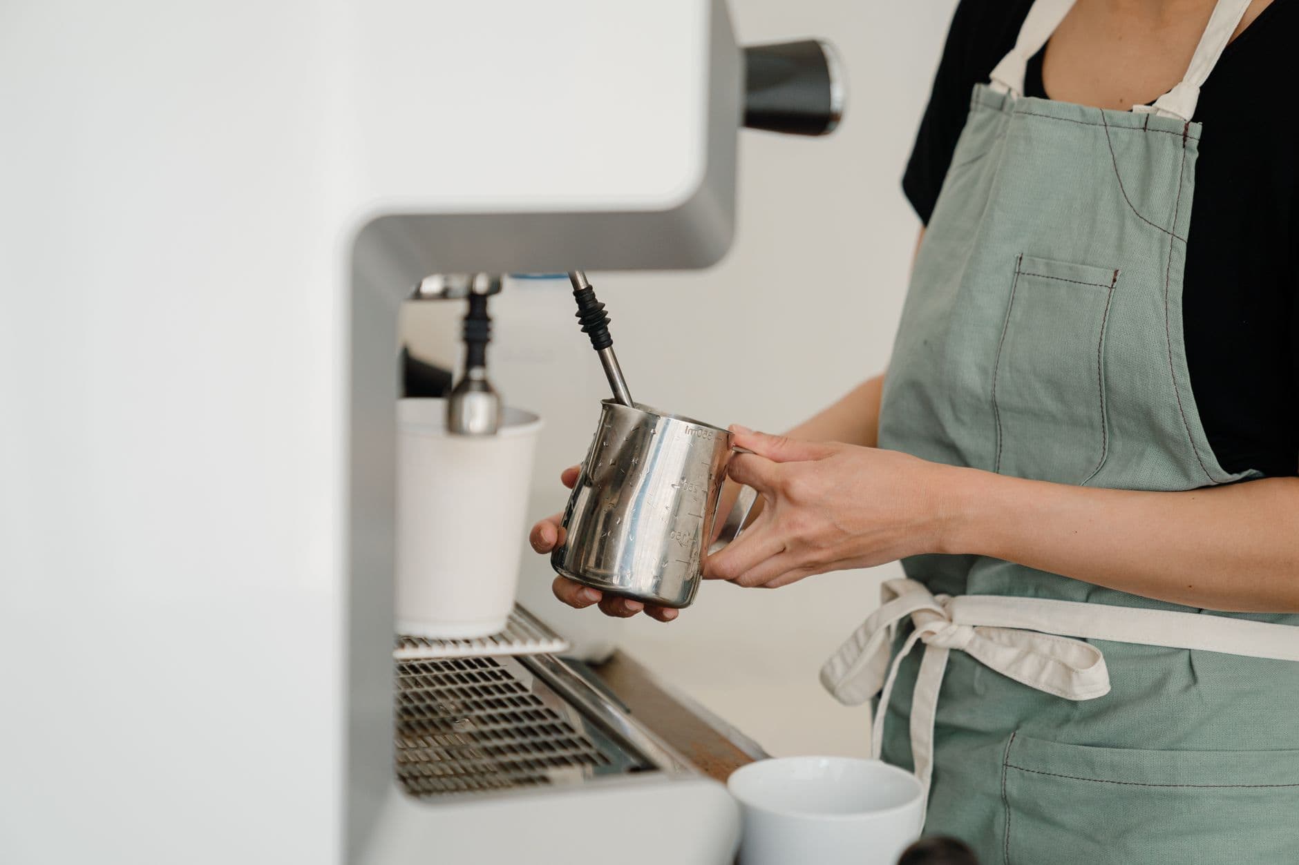 Crop unrecognizable female barista in mint apron uniform whipping milk in stainless steel pitcher using espresso machine