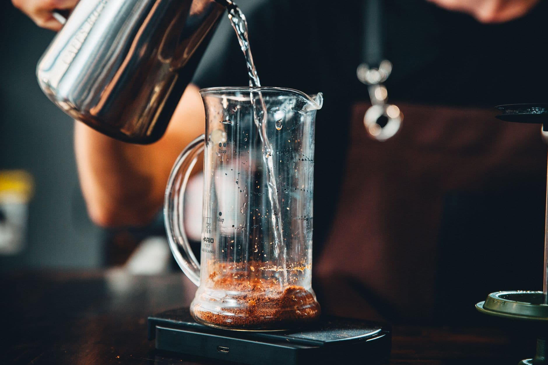 Hands pouring hot water into glass pitcher with coffee grounds for brewing.