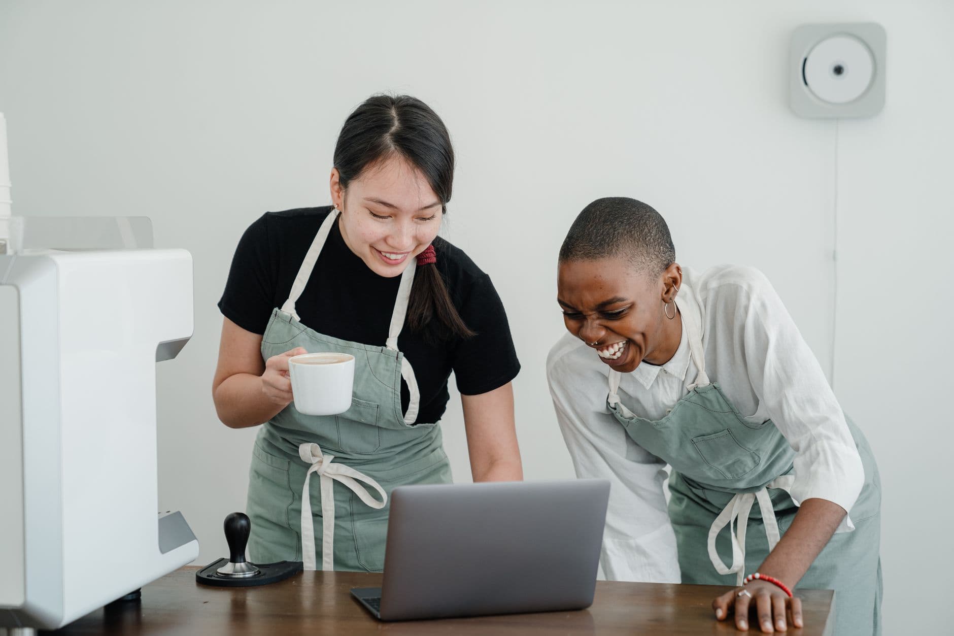 Happy young Asian and African American female coffee house coworkers in aprons laughing while browsing Internet on modern laptop during break