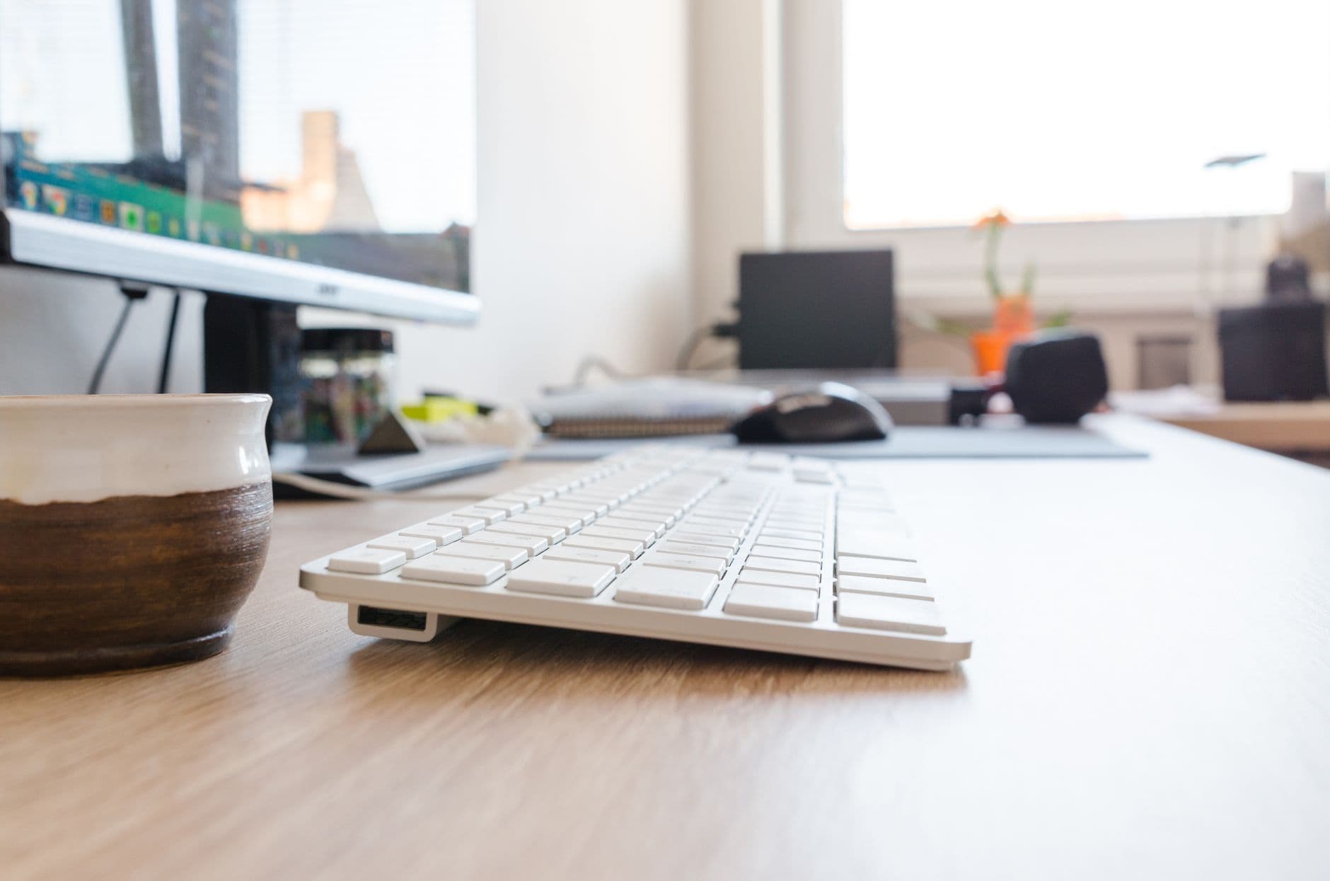 Minimalist office desk setup featuring keyboard, monitor, and coffee cup with natural lighting.