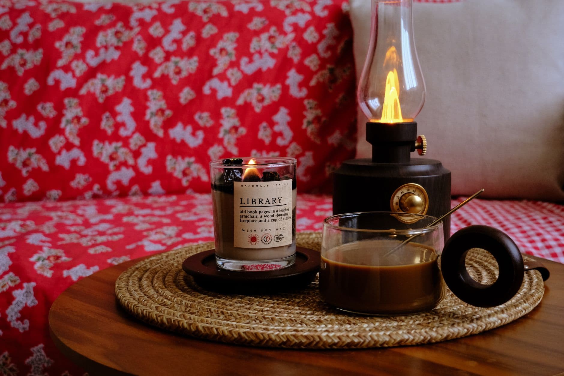 A candle, coffee, and retro lamp create a warm, cozy atmosphere on a wooden table with red patterned fabric.
